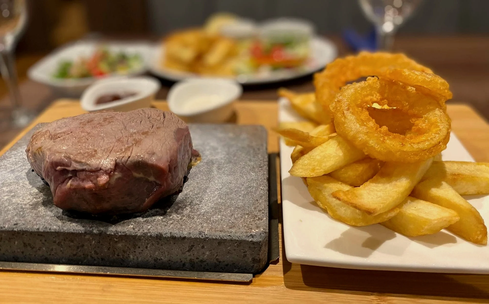Steak on a lava stone next to chips and onion rings