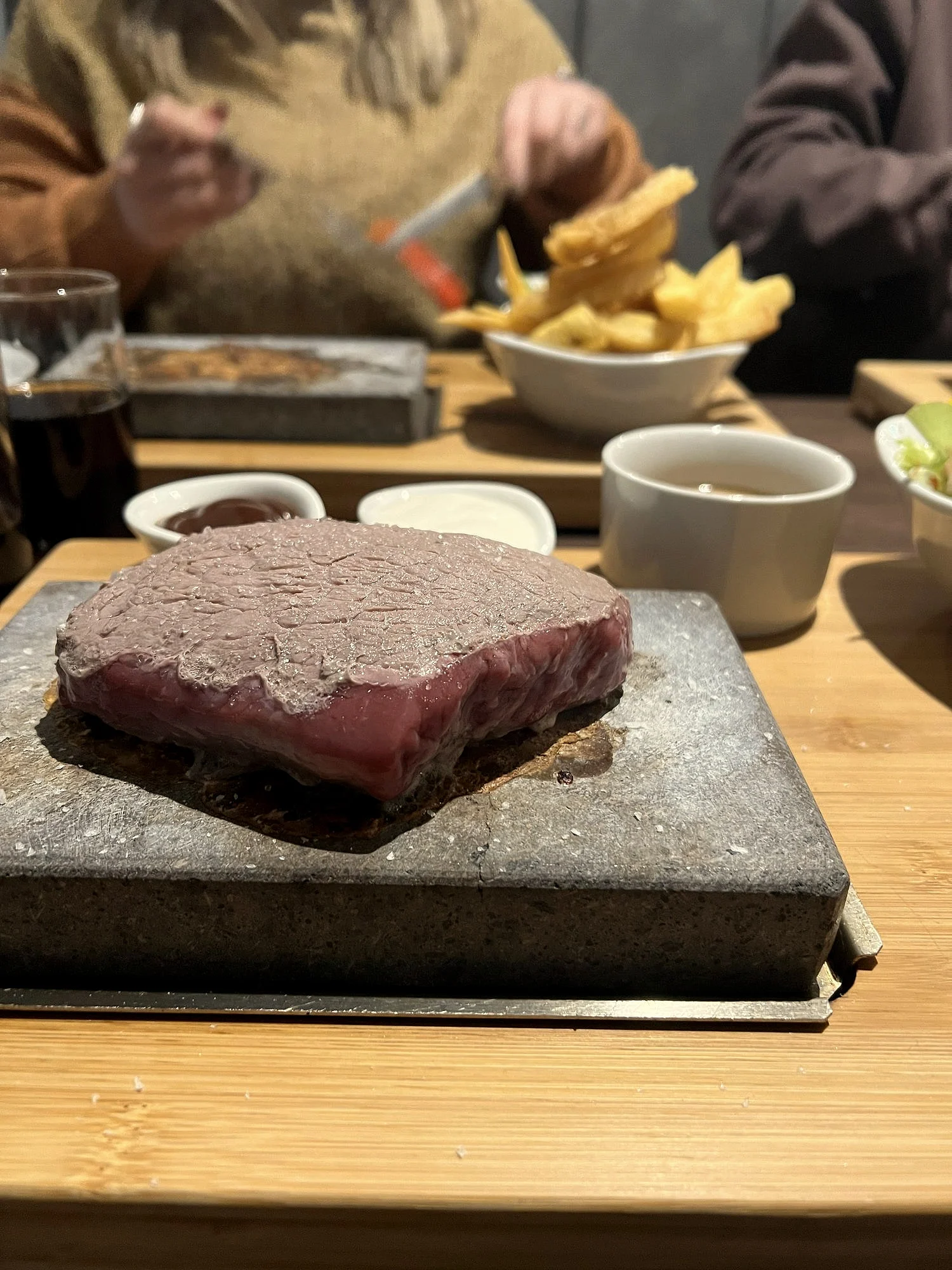 A thick steak on a lava stone, bowls of chips and salad behind