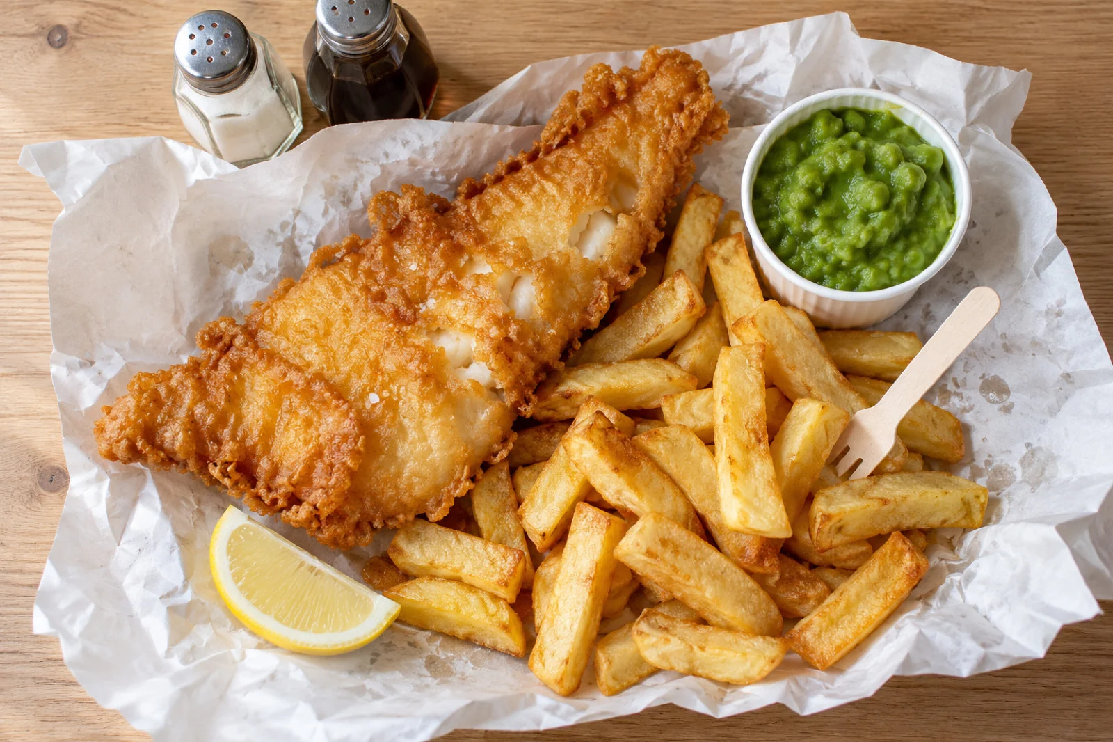 Fish and chips opened out on white paper with mushy peas and a lemon wedge